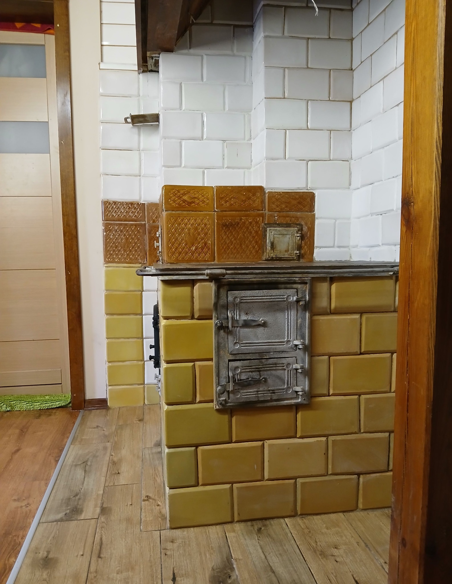 Kuchenny piec kaflowy z metalowymi drzwiczkami, wykończony żółtymi i białymi kaflami, na drewnianej podłodze. Widoczny fragment drzwi i drewniana rama.
