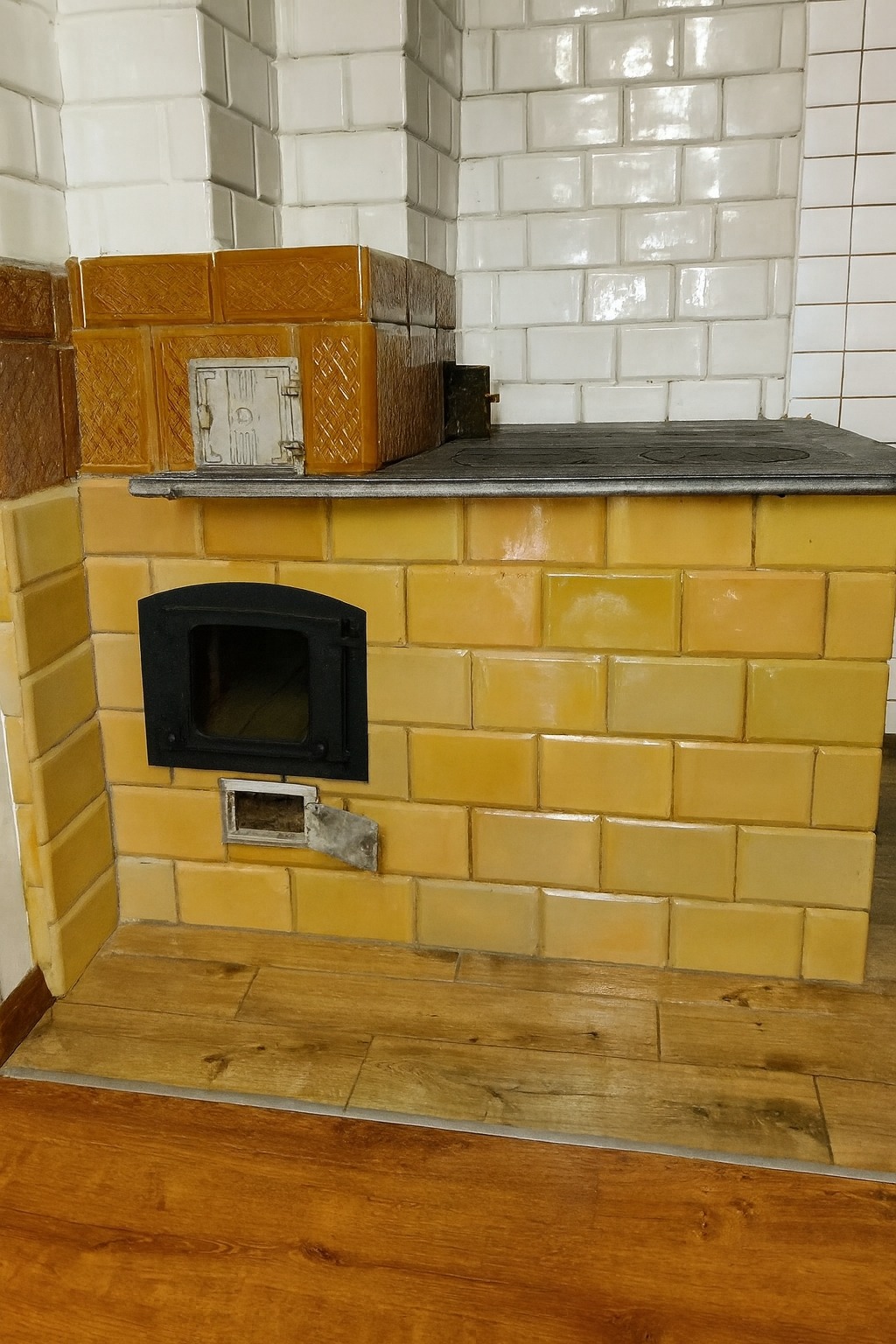 Piec kaflowy w stylu retro z żeliwnym piekarnikiem, zbudowany z selekcjonowanych kafli z odzysku w odcieniach żółci i brązu, na drewnianej podłodze.