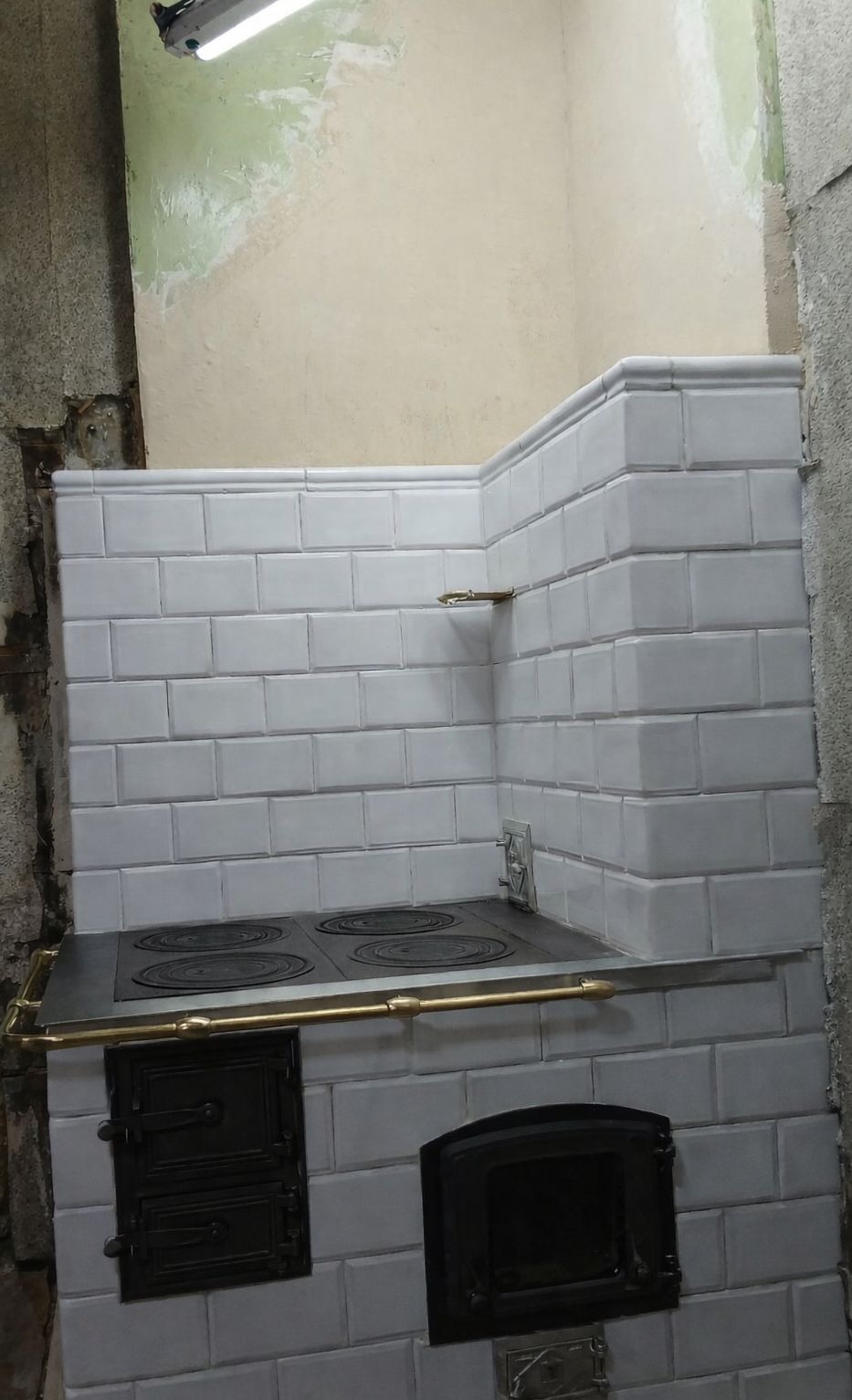 Biały piec kaflowy z czarnymi drzwiczkami i metalowym blatem, wykończony płytkami w stylu retro, w trakcie remontu pomieszczenia. Widoczne surowe ściany.