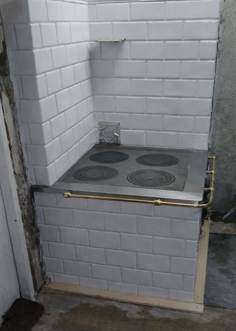 Biały piec kaflowy z czterema palnikami, wykończony złotymi detalami, w trakcie montażu. Widoczne białe płytki i surowy beton w tle.