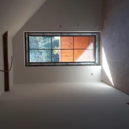 Wnętrze poddasza w trakcie prac wykończeniowych: ściany częściowo pomalowane na biało, surowy beton po prawej, widoczne drewniane belki konstrukcyjne i okno z widokiem na zieleń.