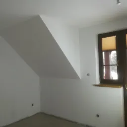 Pomalowane na biało poddasze ze skośnym sufitem, widoczne brązowe okno z plisowanymi żaluzjami i fragment drewnianego domu za oknem.