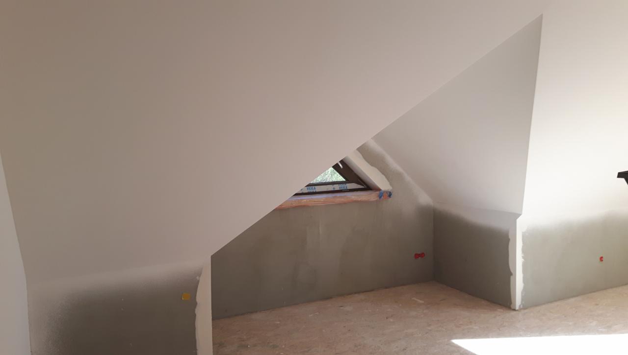 Poddasze po szpachlowaniu i malowaniu, widoczne okno dachowe, ściany w odcieniach szarości i bieli, przygotowane do dalszych prac wykończeniowych, z widocznymi punktami elektrycznymi.