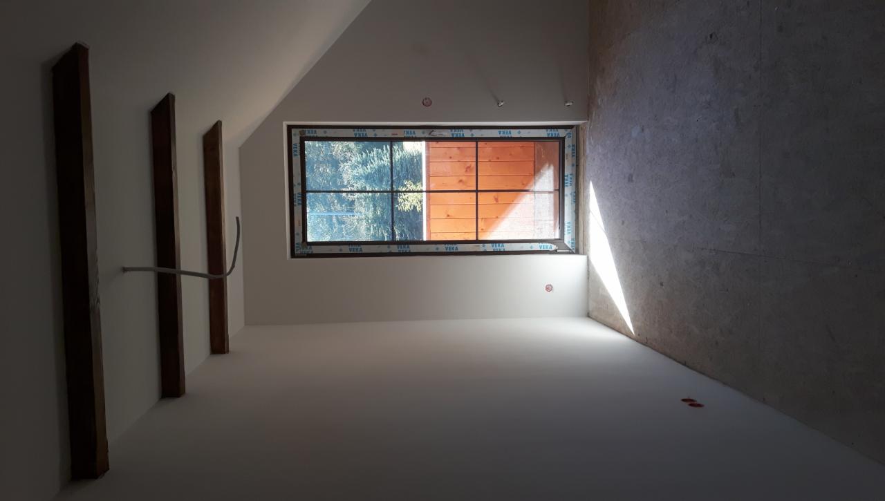 Wnętrze poddasza w trakcie prac wykończeniowych: ściany częściowo pomalowane na biało, surowy beton po prawej, widoczne drewniane belki konstrukcyjne i okno z widokiem na zieleń.