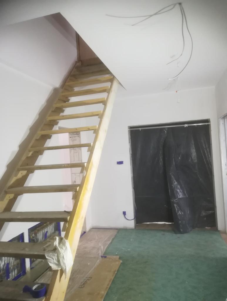 Wnętrze w trakcie remontu: surowe drewniane schody strychowe, zabezpieczona folią podłoga, wystające przewody elektryczne z sufitu, oklejone folią drzwi. Widoczna taśma malarska i karton na podłodze.