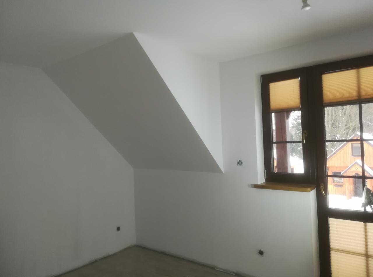 Pomalowane na biało poddasze ze skośnym sufitem, widoczne brązowe okno z plisowanymi żaluzjami i fragment drewnianego domu za oknem.