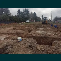 Przygotowany teren pod fundamenty z wykopanymi rowami, widoczny żółty traktor i pracownicy w oddali, w tle drzewa.