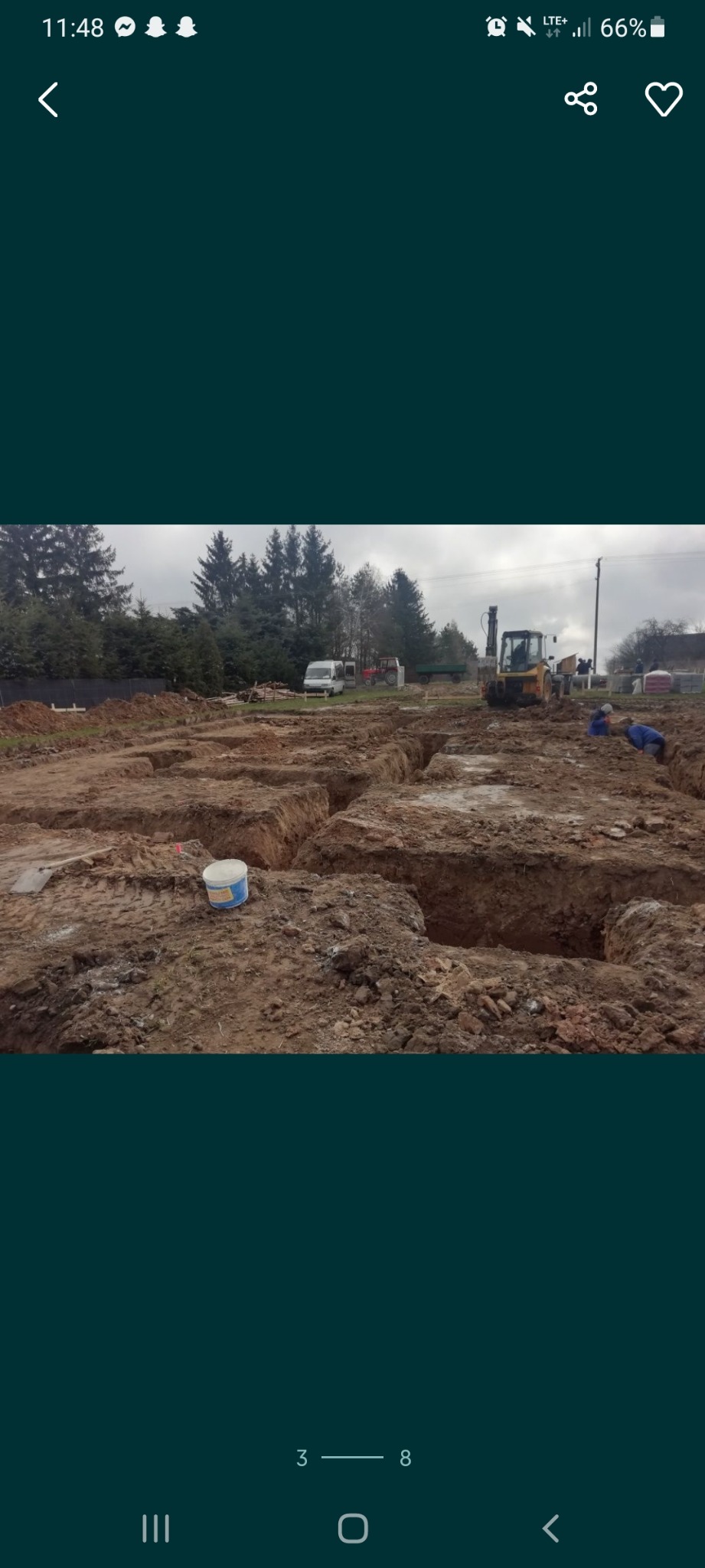 Przygotowany teren pod fundamenty z wykopanymi rowami, widoczny żółty traktor i pracownicy w oddali, w tle drzewa.