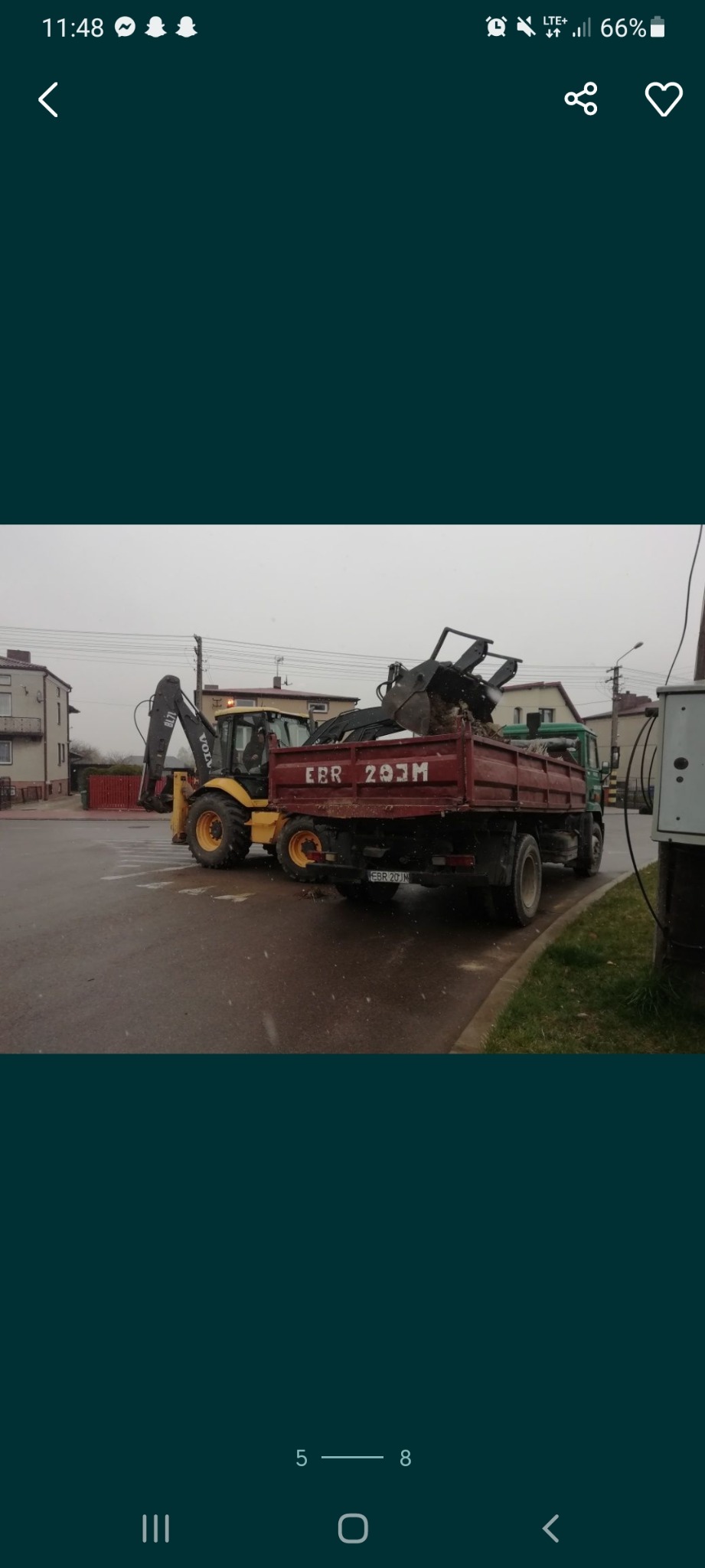 Żółta koparko-ładowarka Volvo BL71 ładuje ziemię do skrzyni ładunkowej czerwonej ciężarówki na tle budynków mieszkalnych