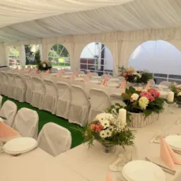 Uroczysty stół w białym namiocie z dekoracjami kwiatowymi, białymi krzesłami z pokrowcami i różowymi serwetkami, przygotowany na przyjęcie.