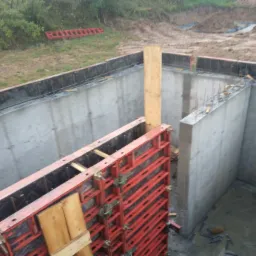 Zalane betonem fundamenty domu w trakcie budowy, widoczne szalunki i wystające pręty zbrojeniowe, w tle teren budowy z zielenią.