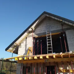 Niedokończony dwukondygnacyjny budynek z pustaków, z widocznym szalunkiem i podporami, drabina oparta o ścianę, błękitne niebo w tle.