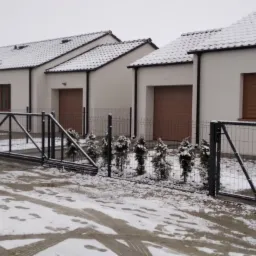 Zimowy krajobraz z nowo wybudowanymi domami jednorodzinnymi, ogrodzonymi szarą, metalową siatką i przesuwną bramą wjazdową, oraz furtką, z małymi, zasypanymi śniegiem tujami wzdłuż ogrodzenia.