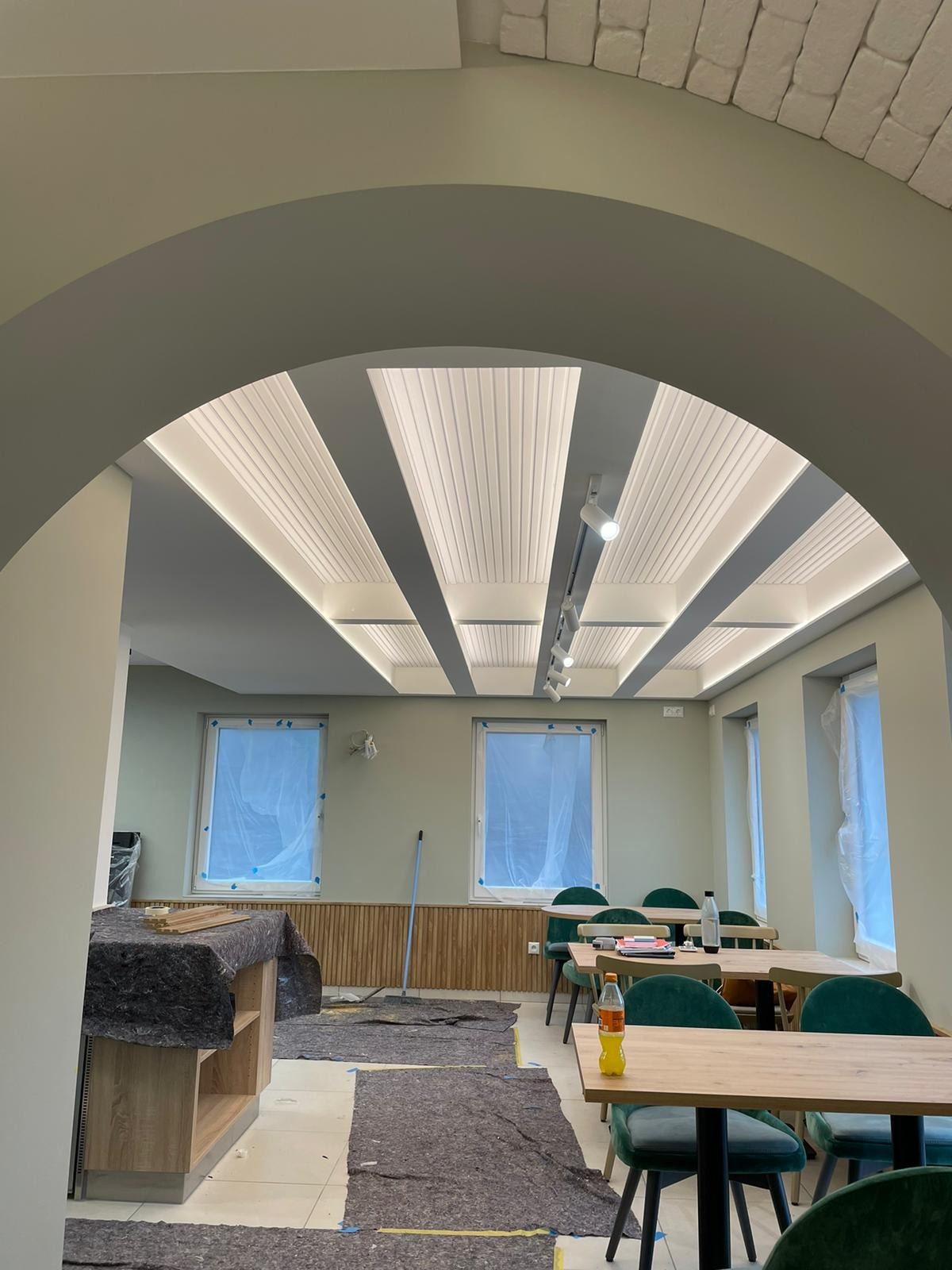 Wnętrze lokalu w trakcie prac wykończeniowych: zabezpieczone okna folią, podłoga przykryta materiałem ochronnym, widoczne oświetlenie sufitowe z paneli i listew LED, świeżo pomalowane ściany...