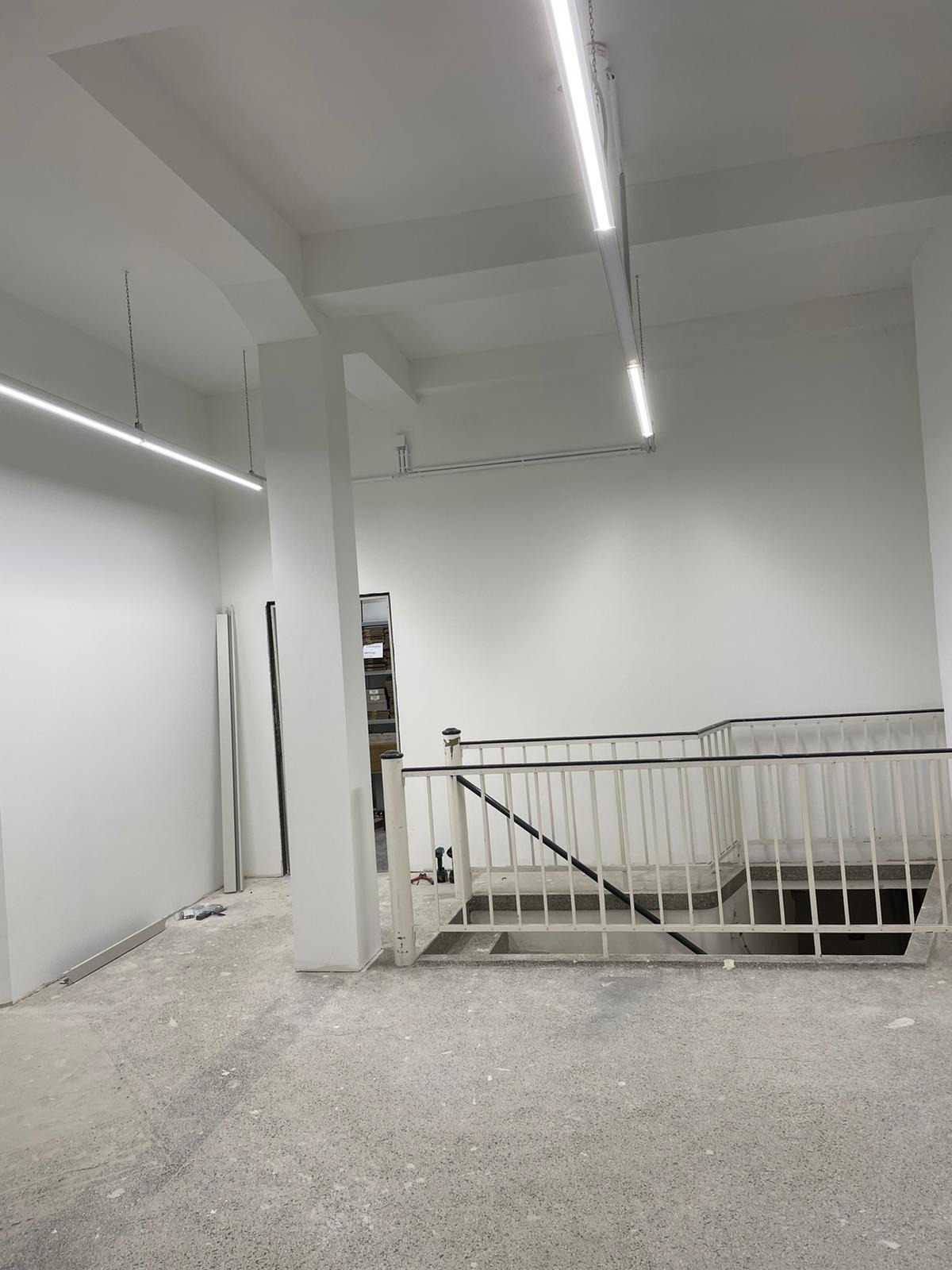 Wnętrze w trakcie wykończenia: białe ściany, betonowa posadzka, balustrada schodowa, oświetlenie liniowe pod sufitem, widoczne ślady prac budowlanych.