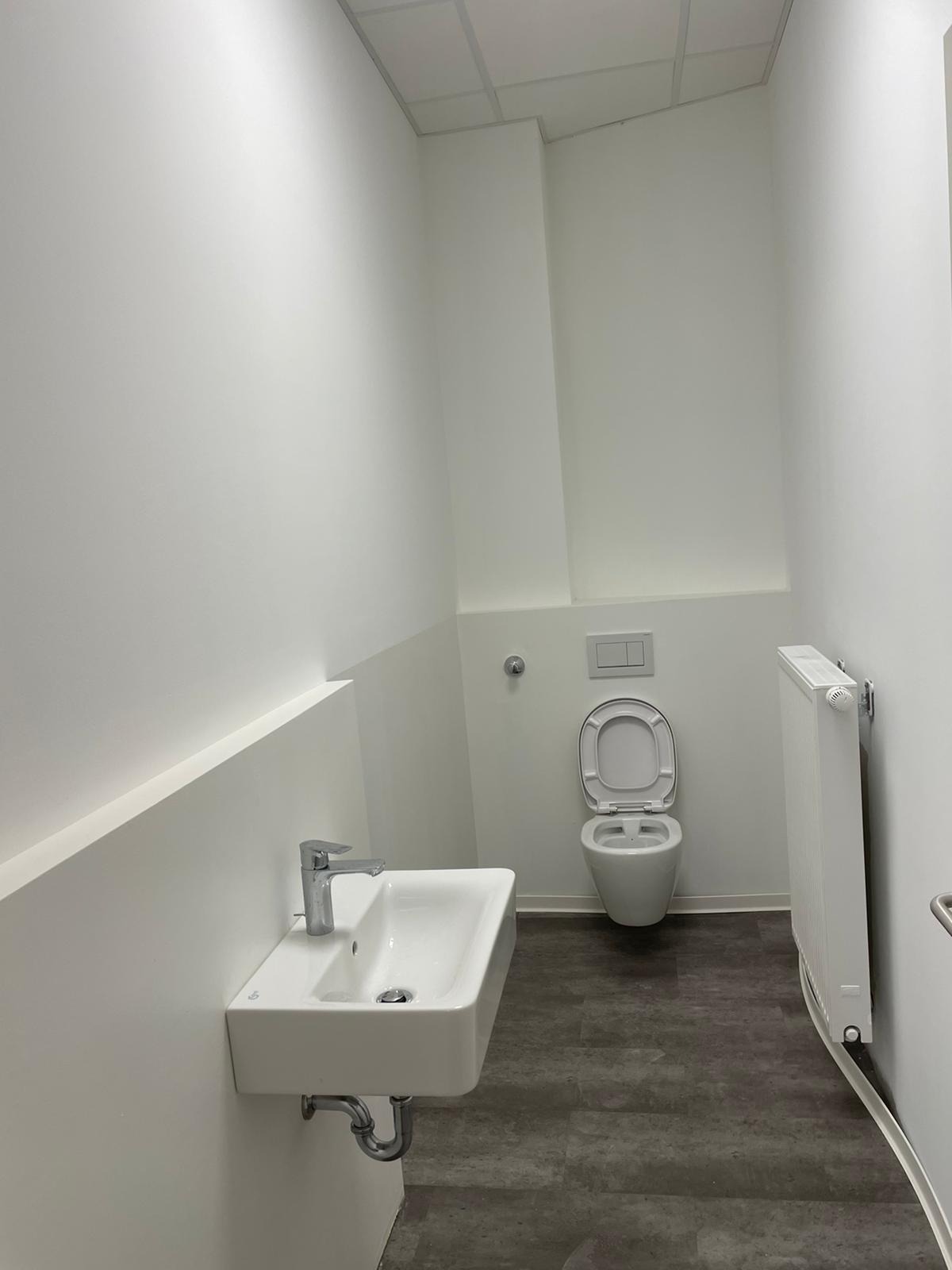 Nowoczesna, minimalistyczna toaleta z umywalką na ścianie, grzejnikiem i otwartą muszlą klozetową, utrzymana w bieli i szarościach.