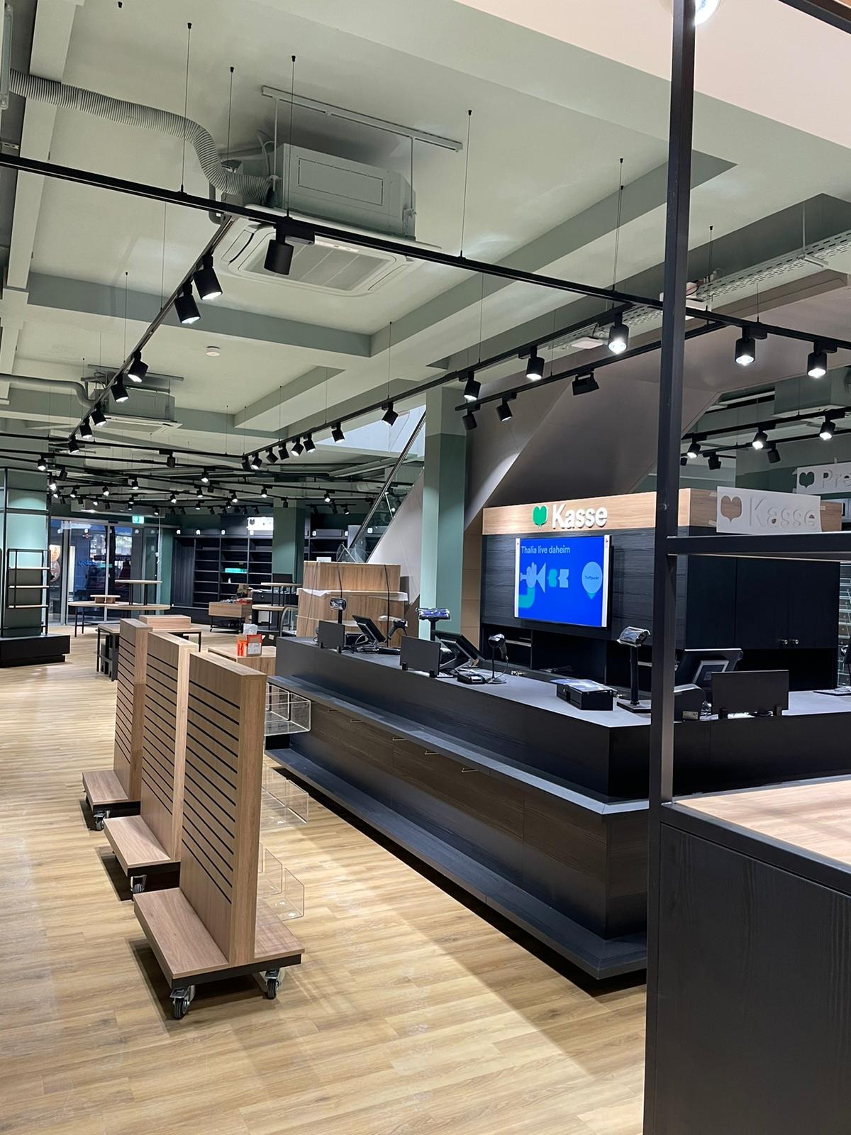 Nowoczesne wnętrze sklepu z kasami w ciemnych kolorach, drewnianą podłogą i oświetleniem punktowym, widoczne regały w tle.