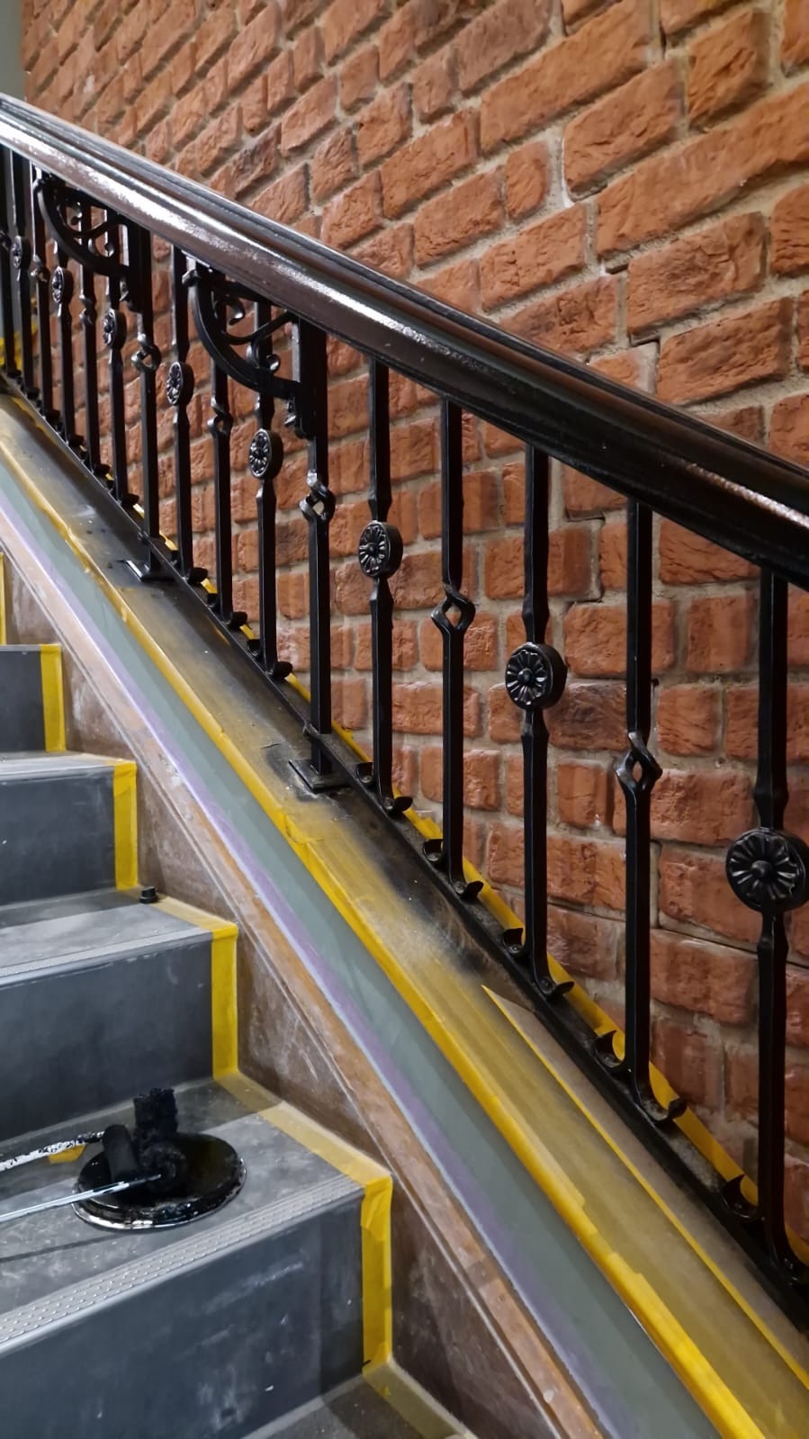Malowanie czarnej, ozdobnej balustrady schodów na tle ceglanej ściany. Schody zabezpieczone taśmą malarską, widoczny wałek i kuweta z farbą.