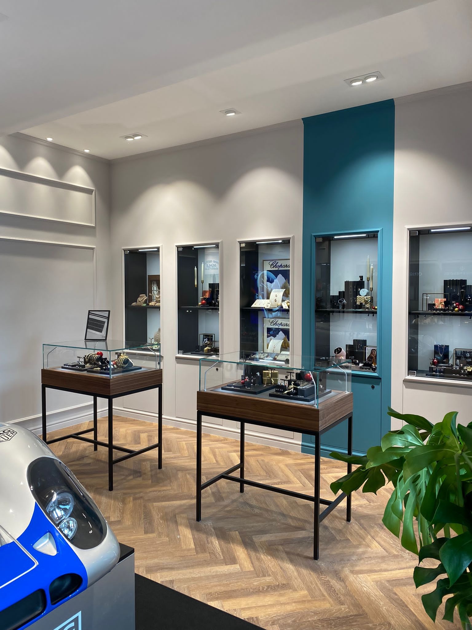 Nowoczesne wnętrze sklepu jubilerskiego z drewnianą podłogą w jodełkę, przeszklonymi witrynami na czarnych nóżkach eksponującymi biżuterię i zegarki, oraz witrynami ściennymi z oświetleniem LED...