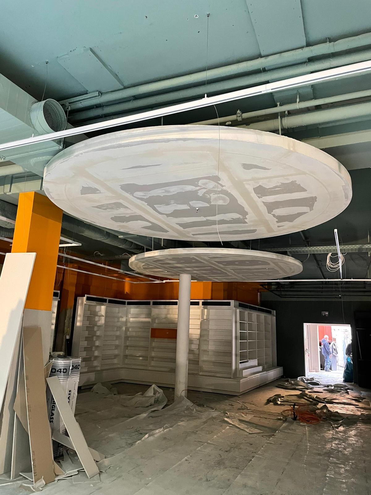 Dwa okrągłe podwieszane sufity z płyt kartonowo-gipsowych w trakcie szpachlowania, widoczne instalacje sufitowe i pomarańczowe elementy konstrukcyjne, podłoga zabezpieczona folią, w tle białe regały.