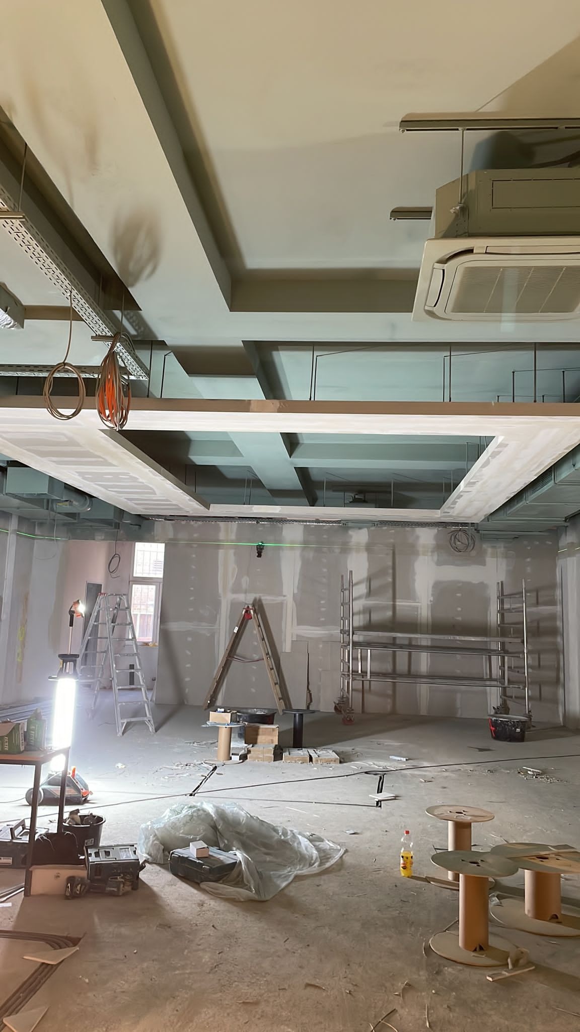 Wnętrze w trakcie prac wykończeniowych: widoczne profile sufitowe, instalacje wentylacyjne, klimatyzacja, ściany przygotowane pod malowanie, drabiny, rusztowania i narzędzia.