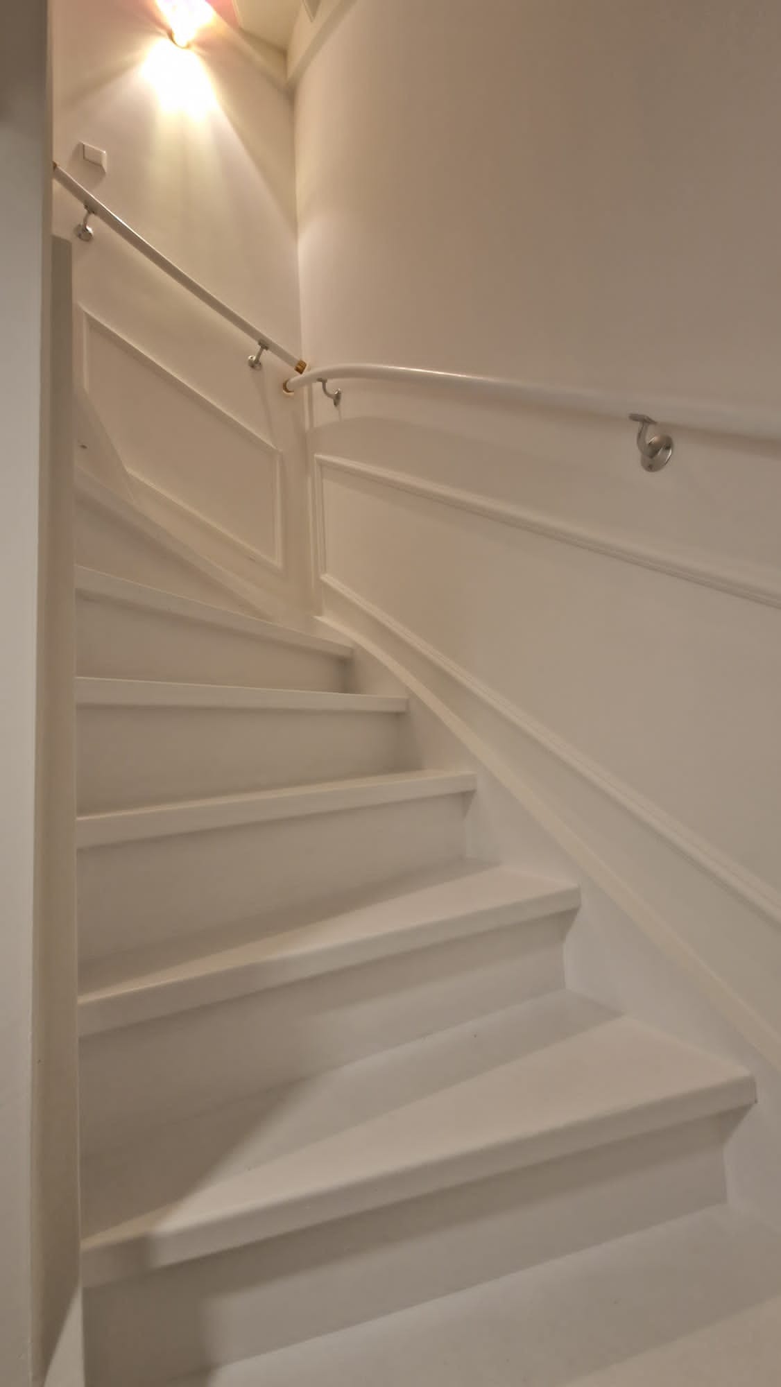 Białe schody z ozdobnymi panelami ściennymi i poręczą, oświetlone od góry.