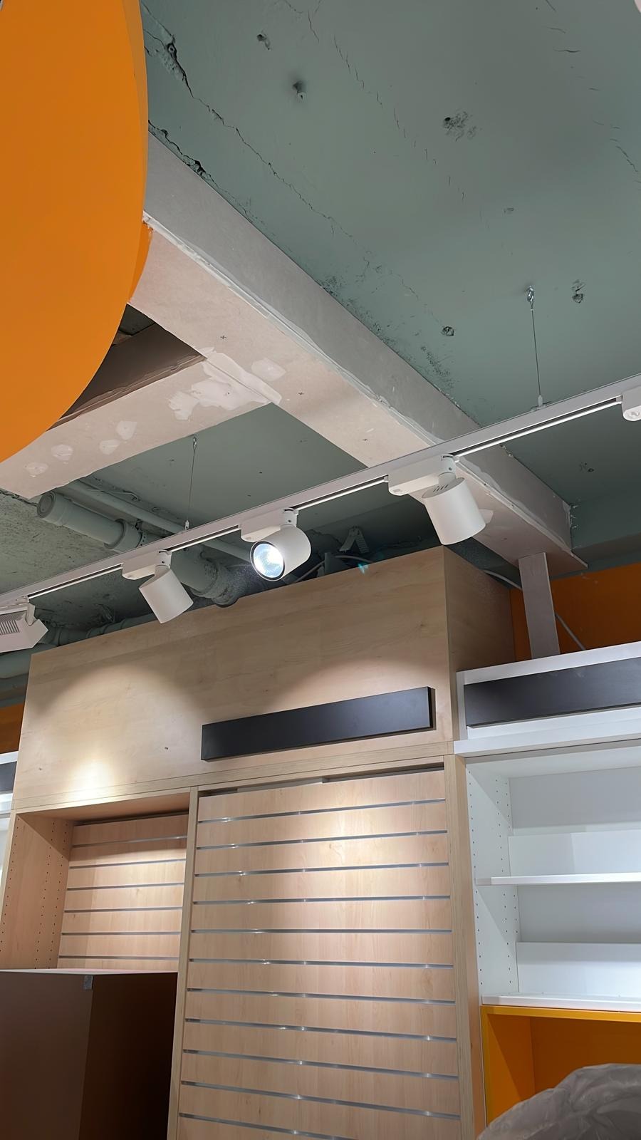 Wnętrze sklepu w trakcie wykańczania, widoczne oświetlenie szynowe, drewniane panele ścienne z metalowymi listwami i sufit z widocznymi pęknięciami.