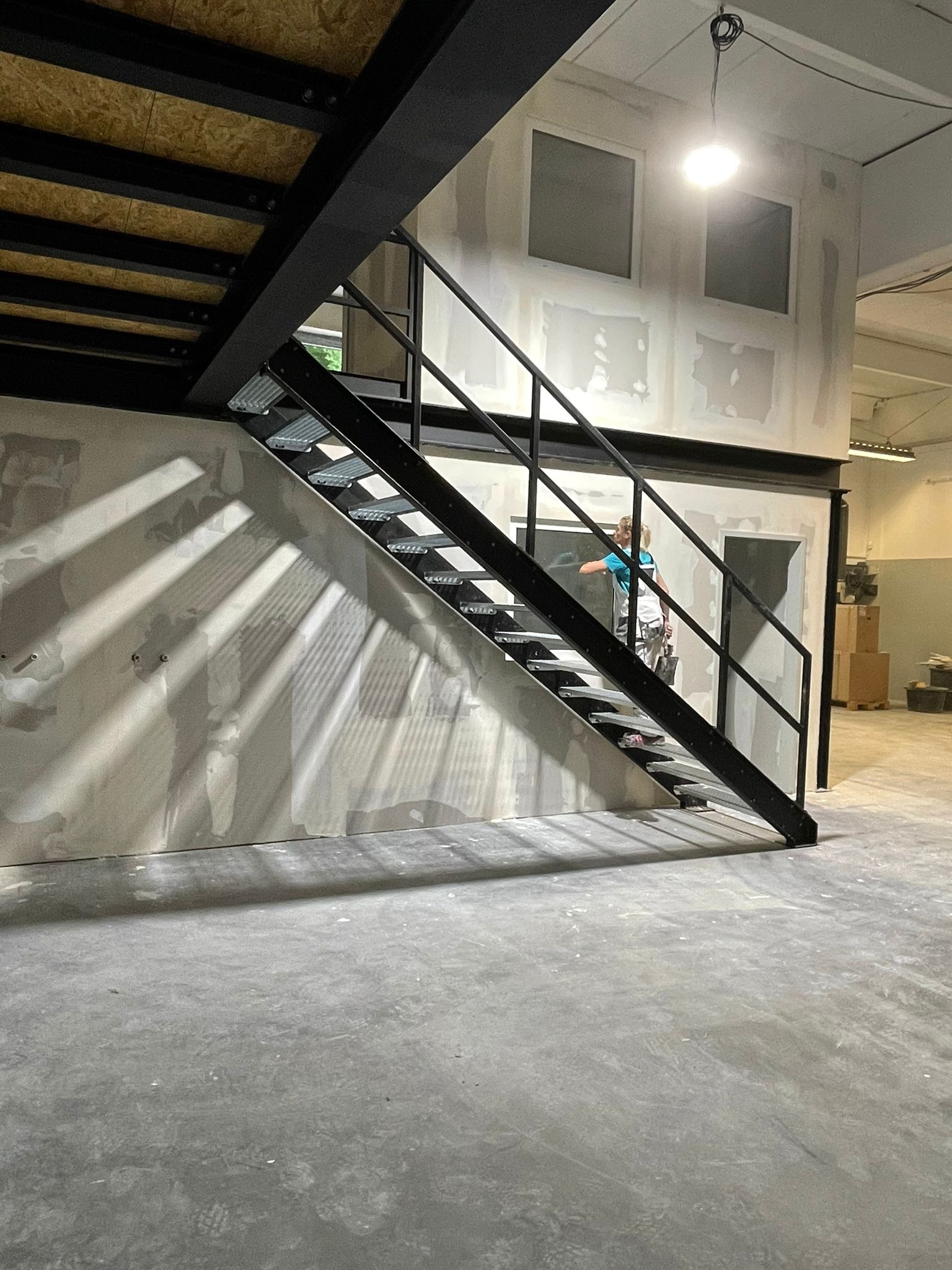 Wnętrze w trakcie prac wykończeniowych: metalowe schody z ażurowymi stopniami, częściowo wyszpachlowane ściany, widoczne ślady po łatach, osoba w stroju roboczym na schodach, surowa betonowa podłoga.