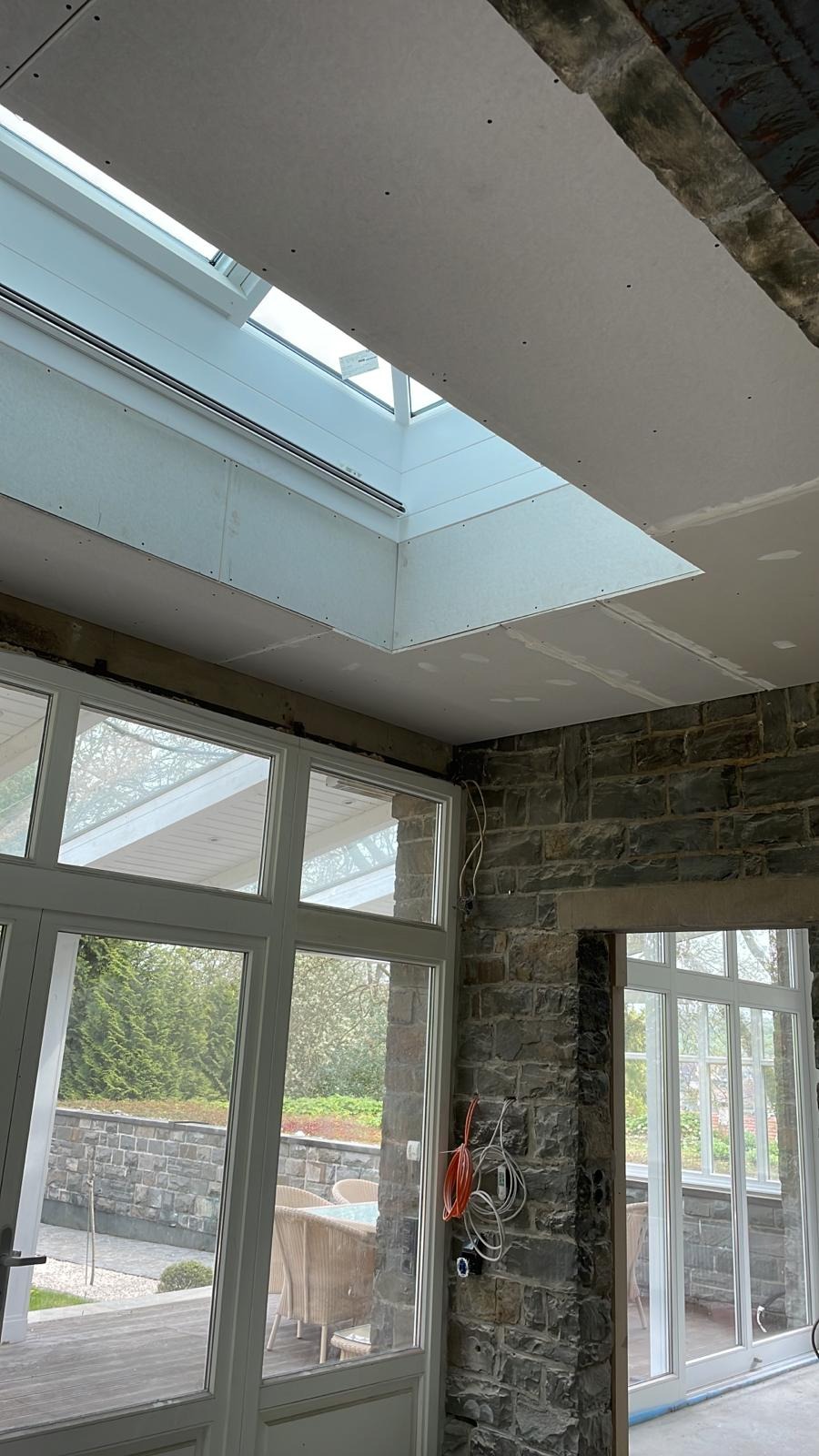 Wnętrze w trakcie remontu: montaż płyty karton gips na suficie z oknem dachowym, widoczne ściany z kamienia i duże okna tarasowe.