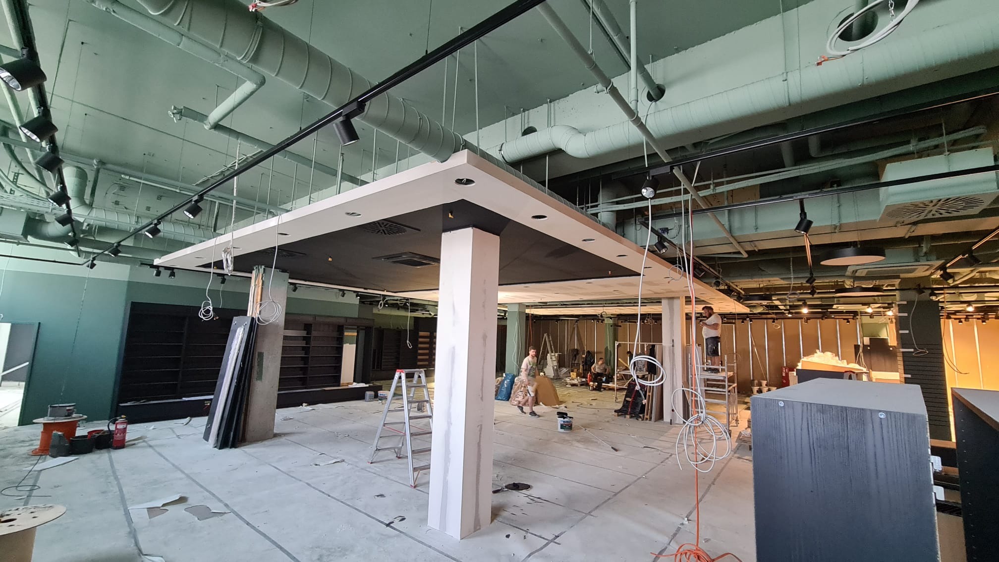 Wnętrze sklepu w trakcie wykańczania, widoczne instalacje pod sufitem, regały i pracownicy na drabinach.