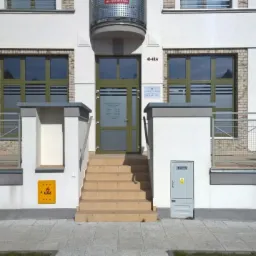 Wejście do budynku z oznaczeniem numeru 41-42a, schody z jasnych płytek prowadzą do przeszklonych drzwi z widoczną tabliczką informacyjną, okna z zielonymi ramami, żółta tabliczka z napisem GAZ.