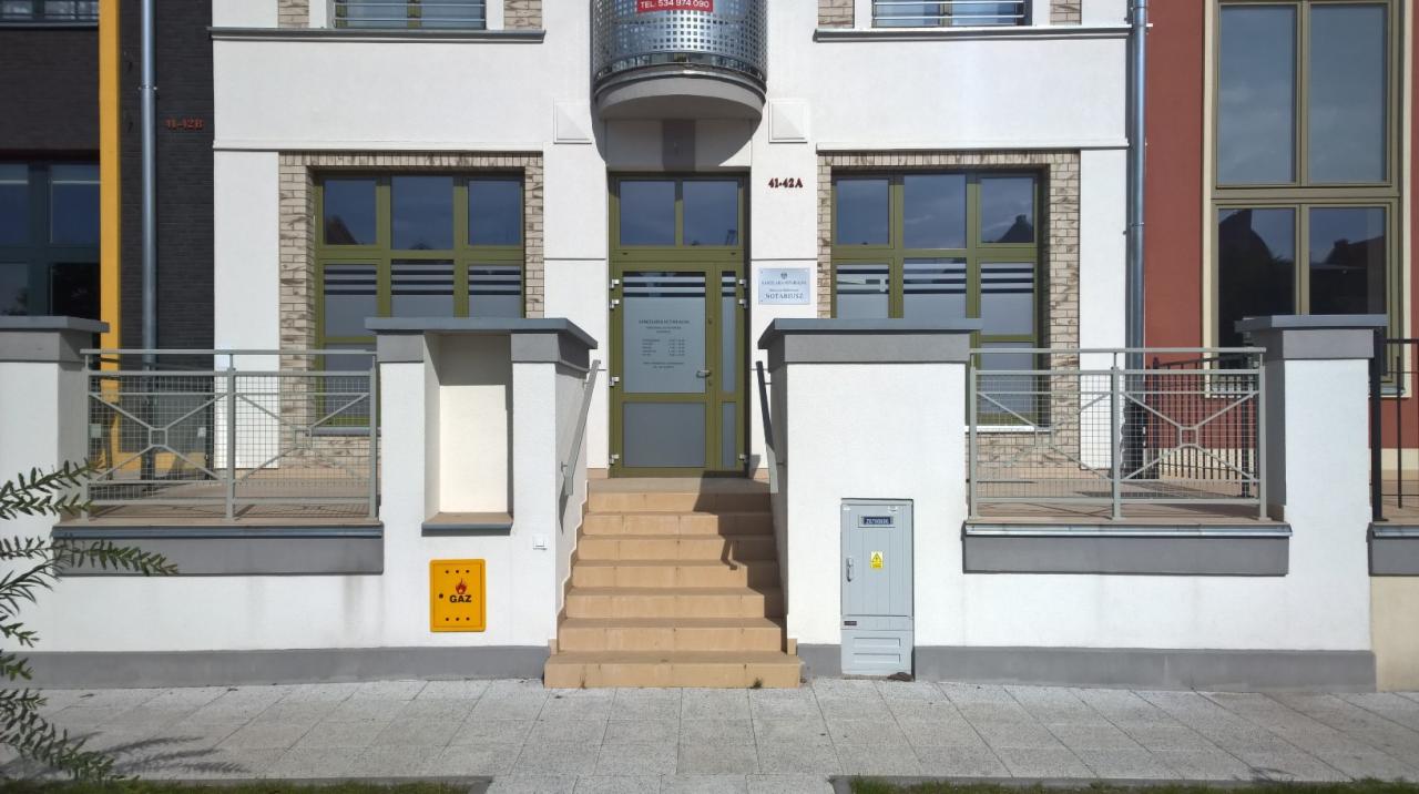 Wejście do budynku z oznaczeniem numeru 41-42a, schody z jasnych płytek prowadzą do przeszklonych drzwi z widoczną tabliczką informacyjną, okna z zielonymi ramami, żółta tabliczka z napisem GAZ.
