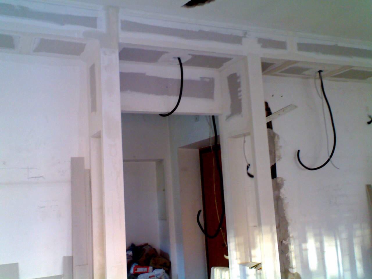 Wnętrze w trakcie prac wykończeniowych: widoczne profile pod płyty gipsowo-kartonowe na suficie i ścianach, wiszące kable elektryczne, fragment drzwi w tle.