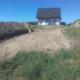 Nowo wybudowany, parterowy dom z szarą dachówką w Warszawie, otoczony usypaną ziemią i trawą, w słoneczny dzień.