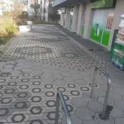 Ułożona kostka brukowa w kształcie heksagonów przed budynkiem sklepu Żabka, widoczne stojaki na rowery i budynek mieszkalny w tle.