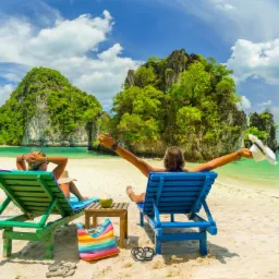 Dwie osoby relaksują się na plaży na leżakach z widokiem na turkusową wodę i formacje skalne porośnięte zielenią; jedna osoba trzyma kapelusz w górze, obok stolik z kokosem, kolorowa torba plażowa...