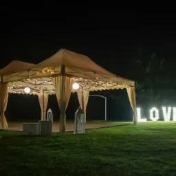 Dwa beżowe namioty z białymi zasłonami, oświetlone girlandami i kulami, drewniany parkiet, białe lampiony i skrzynki, napis LOVE z podświetlanych liter na trawie, nocne oświetlenie.