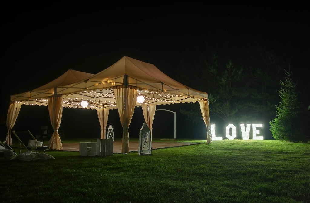Dwa beżowe namioty z białymi zasłonami, oświetlone girlandami i kulami, drewniany parkiet, białe lampiony i skrzynki, napis LOVE z podświetlanych liter na trawie, nocne oświetlenie.