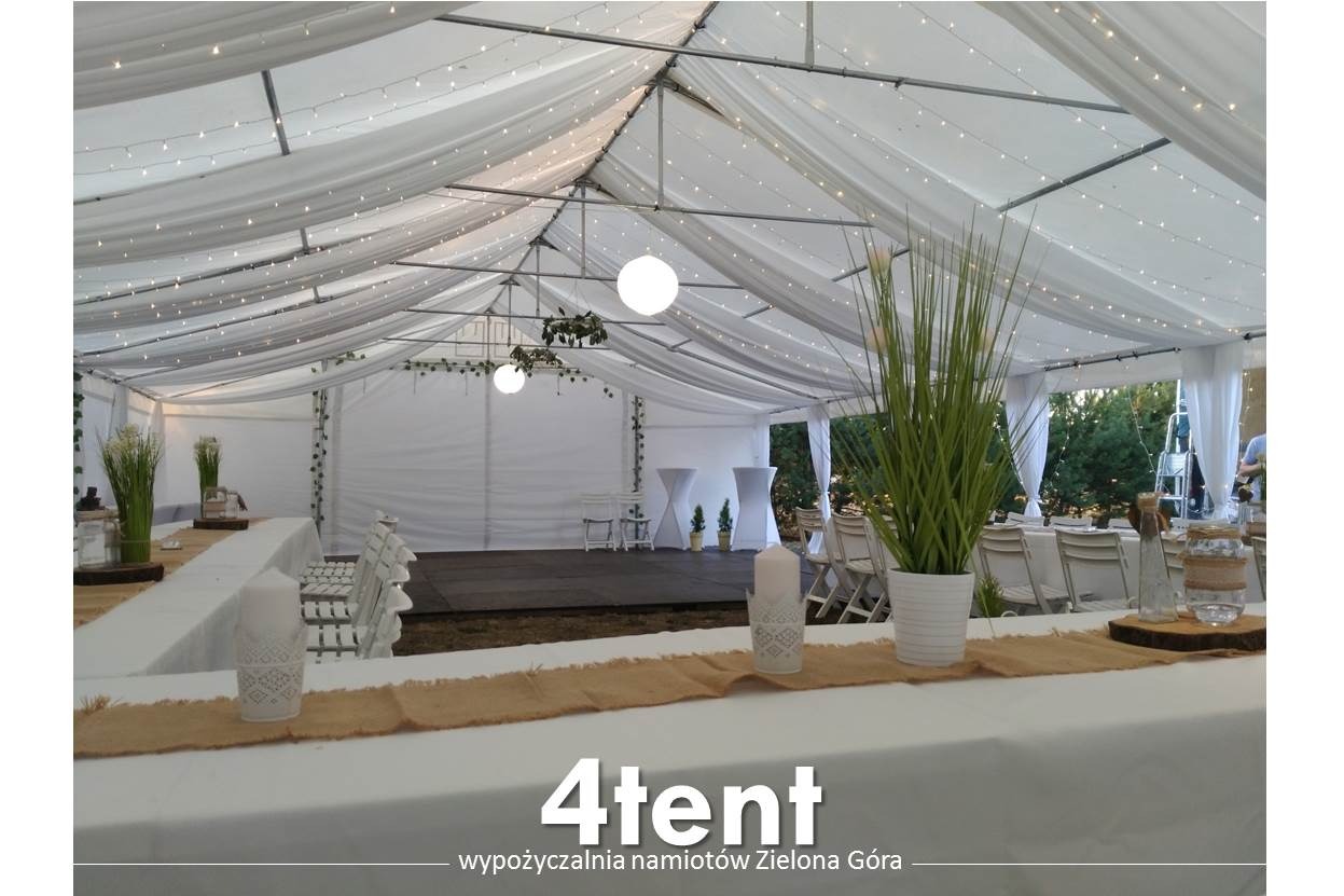 Wnętrze białego namiotu z dekoracjami: girlandy świetlne, wiszące kule, rośliny w doniczkach, białe krzesła i stoły z jutowymi bieżnikami.