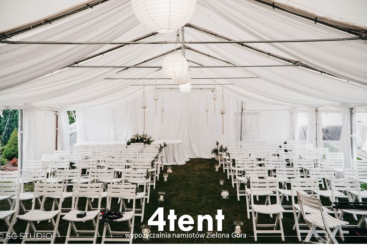 Biały namiot z rozstawionymi rzędami białych krzeseł, dekoracjami kwiatowymi i lampionami, przygotowany na uroczystość.