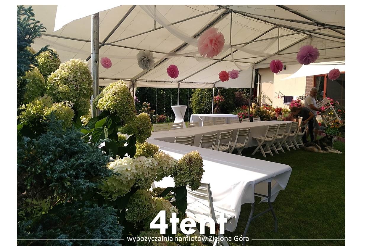 Biały namiot imprezowy z dekoracjami z papierowych kul, ustawione białe stoły i krzesła, na pierwszym planie hortensje, w tle osoby przygotowujące przestrzeń i pies.