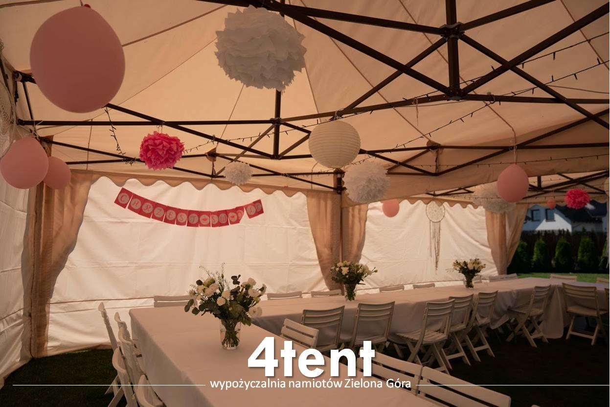 Udekorowany namiot z rozłożonymi stołami, kwiatami w wazonach, balonami i papierowymi ozdobami wiszącymi pod sufitem, przygotowany na uroczystość. Widoczny baner z napisem 'Chrzest'.