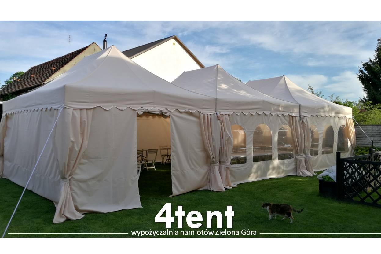 Rozłożony, beżowy namiot modułowy z dekoracyjnymi falbanami na obrzeżach, z widocznymi krzesłami wewnątrz, stojący na trawie, z kotem spacerującym obok.