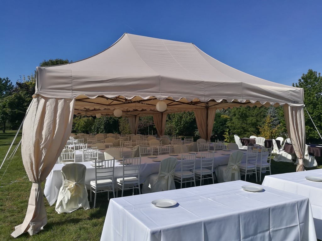 Beżowy namiot plenerowy z białymi stołami i krzesłami, przygotowany na uroczystość na trawniku w słoneczny dzień.