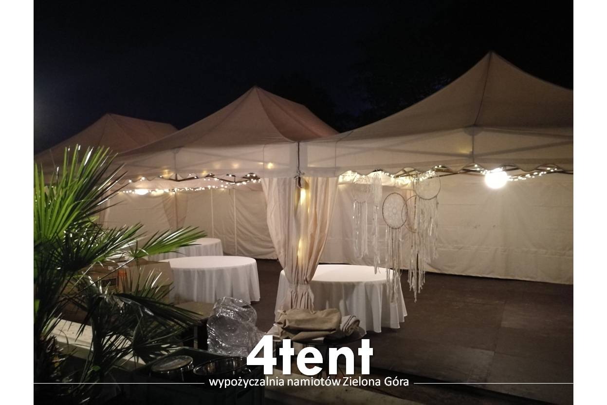 Udekorowane namioty z oświetleniem i łapaczami snów, białe okrągłe stoły, rośliny doniczkowe w tle nocnego nieba.