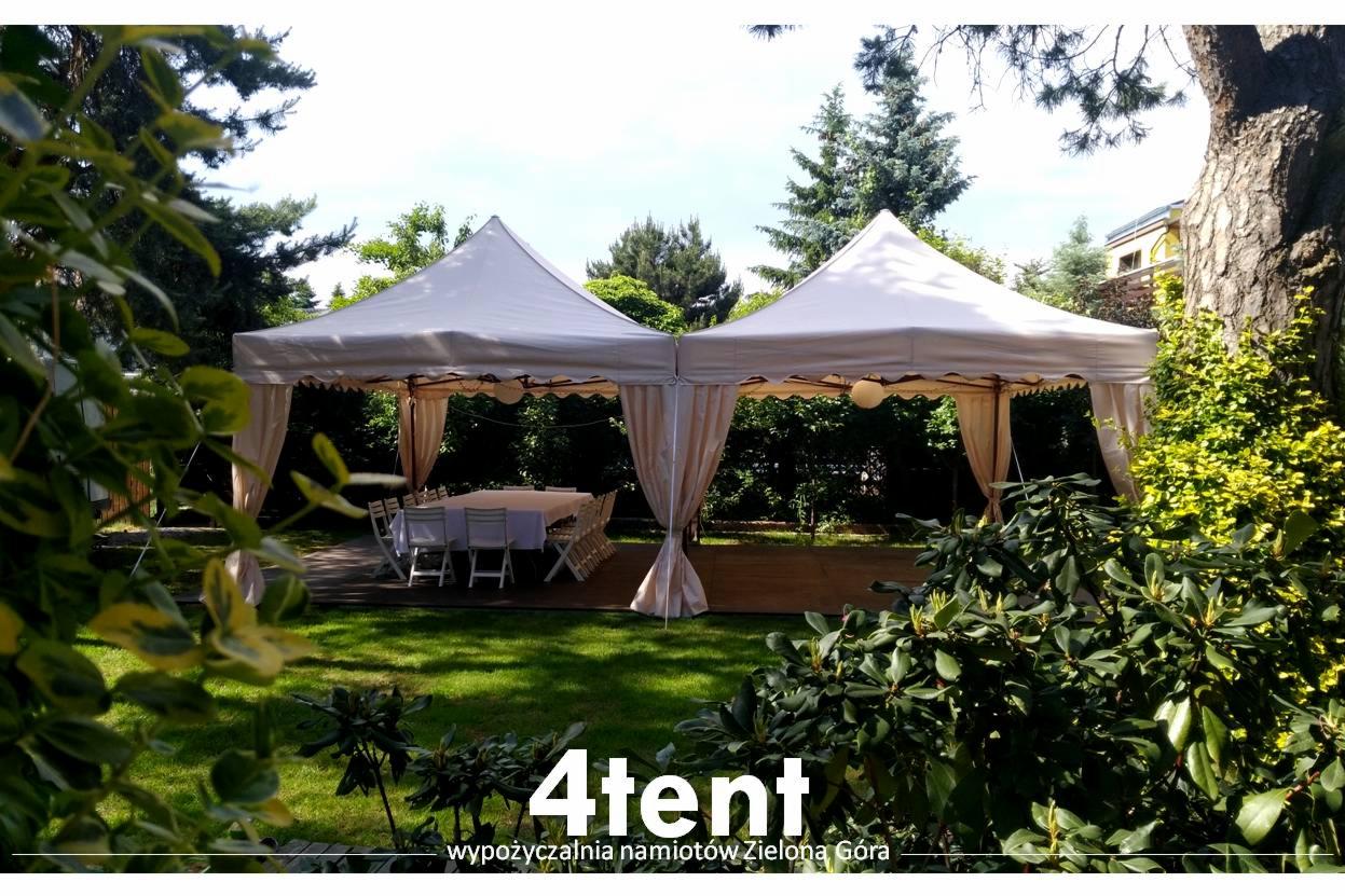 Dwa białe namioty połączone ze sobą, z beżowymi zasłonami, ustawione na drewnianym podeście w ogrodzie, stół i krzesła wewnątrz namiotu.