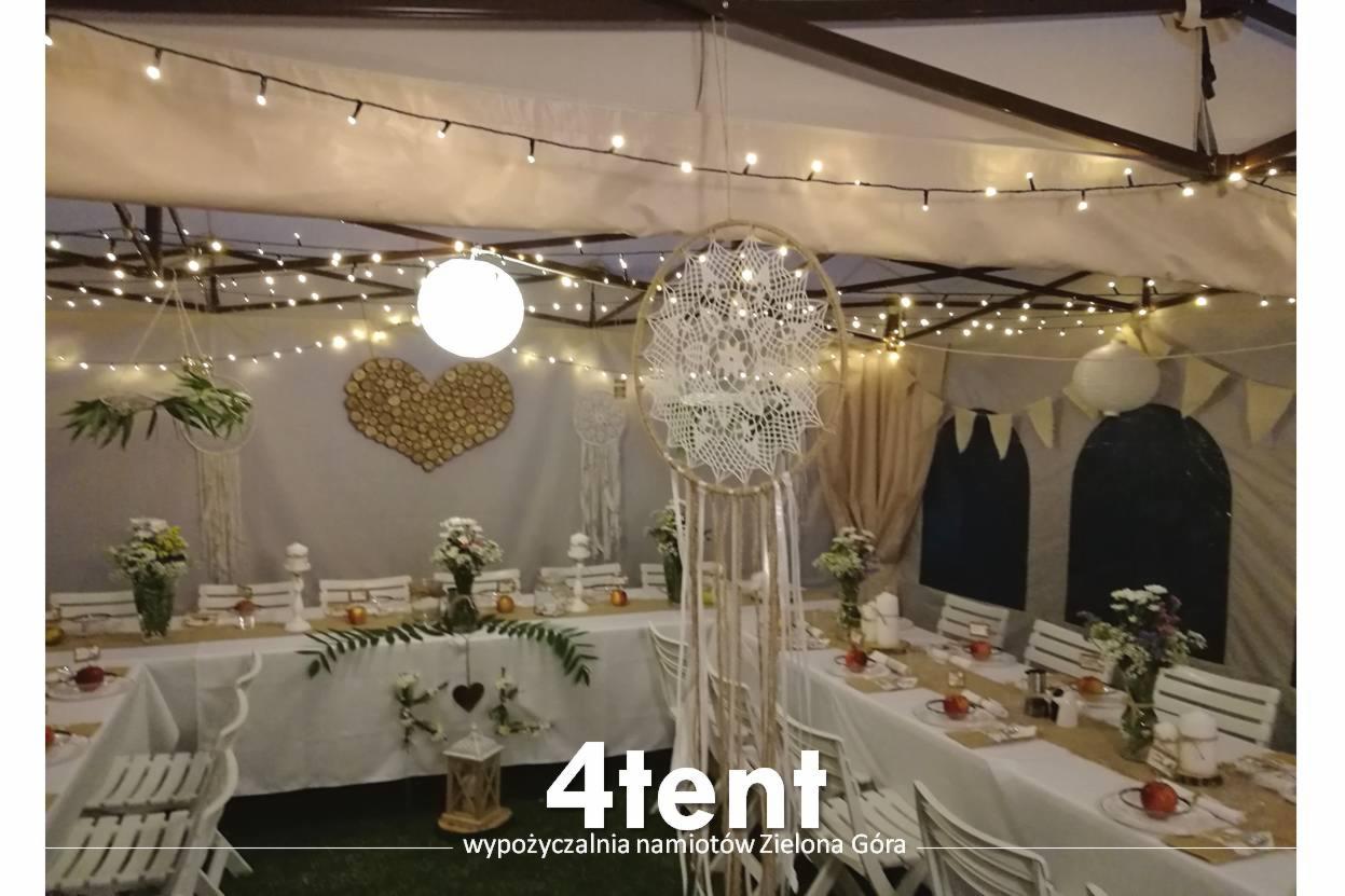 Dekoracja wnętrza namiotu z drewnianym sercem, łapaczami snów, kwiatami, girlandami świetlnymi i białymi krzesłami, przygotowanego na imprezę.