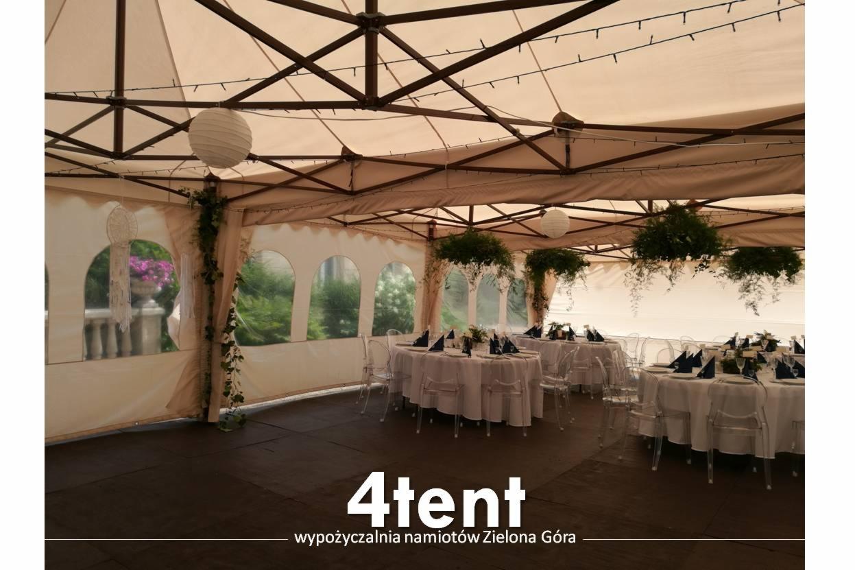 Udekorowany namiot bankietowy z okrągłymi stołami i przezroczystymi krzesłami, oświetlony lampionami i ozdobiony zielenią, Zielona Góra.