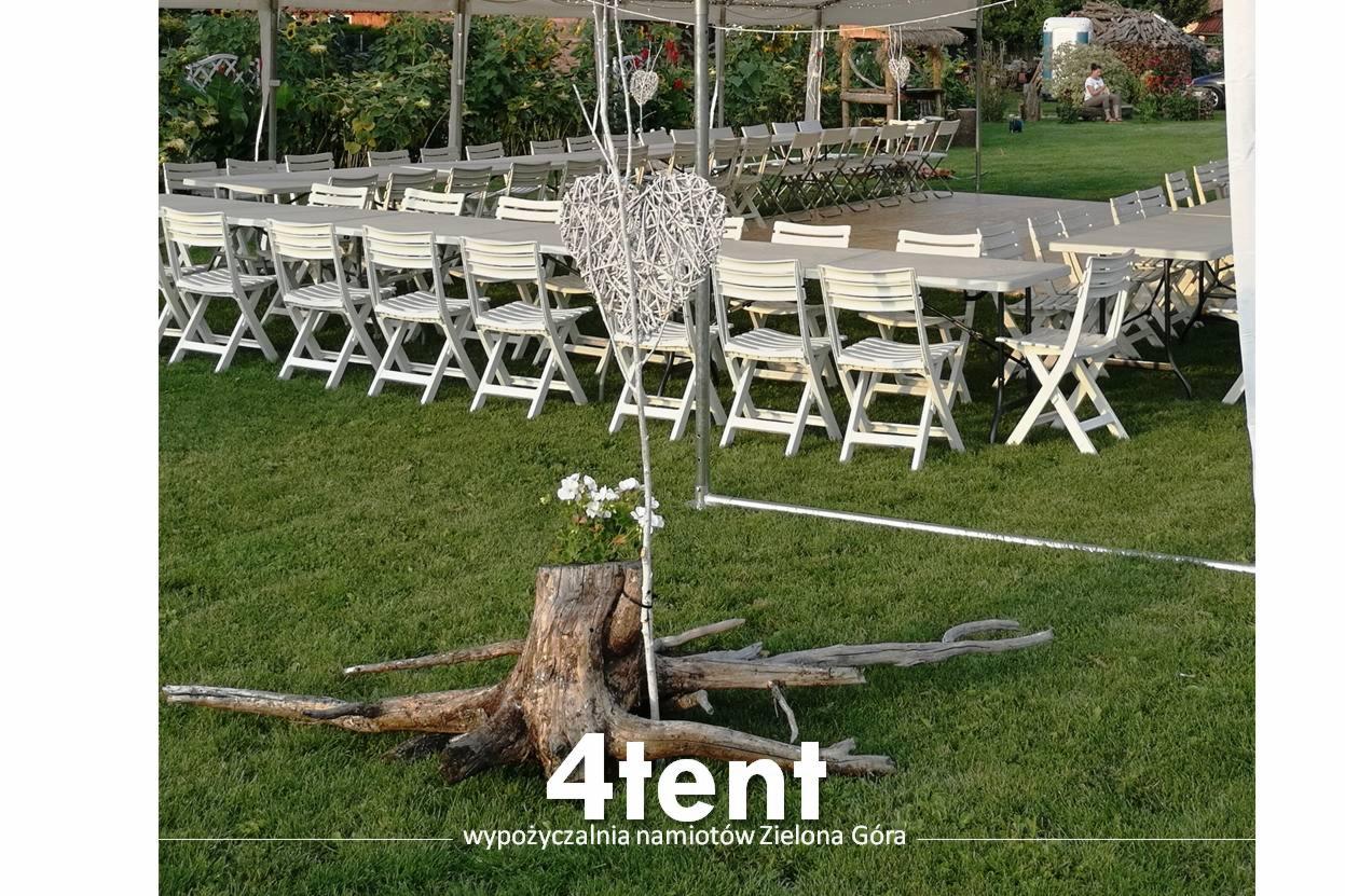 Ustawione stoły i krzesła pod białym namiotem na zielonym trawniku, dekoracje w postaci wiklinowych serc i pnia drzewa z kwiatami. Widoczny napis '4tent wypożyczalnia namiotów Zielona Góra'.