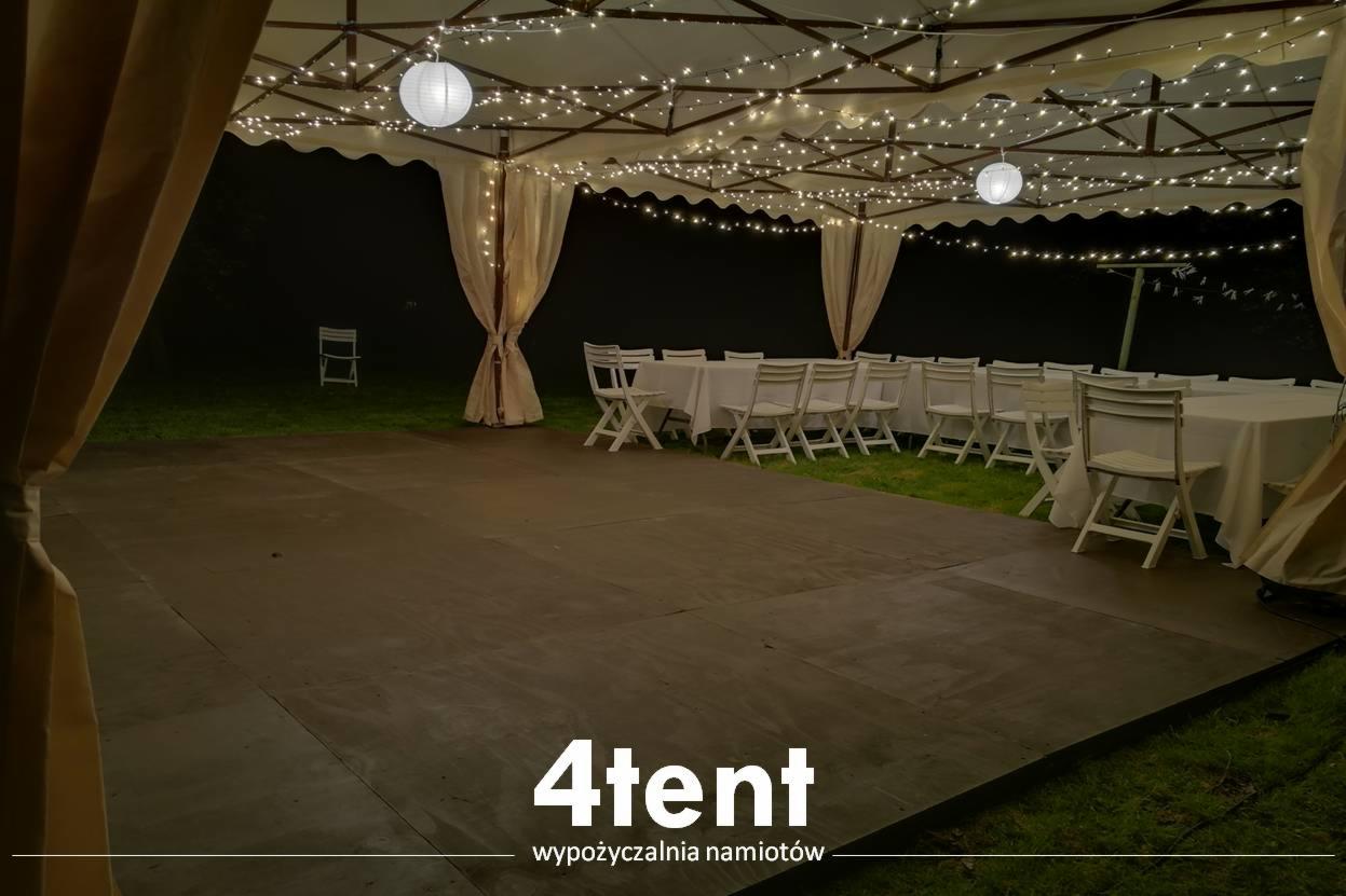 Udekorowany namiot imprezowy z białymi krzesłami i stołami, oświetlony girlandami świetlnymi i papierowymi lampionami, widok na parkiet i zieleń w tle, logo firmy w dolnej części zdjęcia.